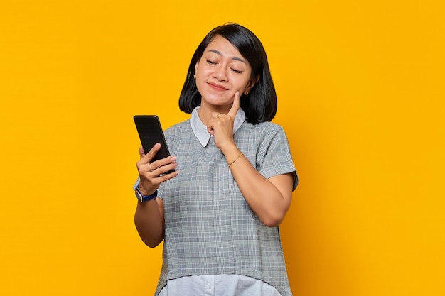 Sonriente joven mujer asiática pensando en pregunta con el dedo en la barbilla y sosteniendo el teléfono móvil