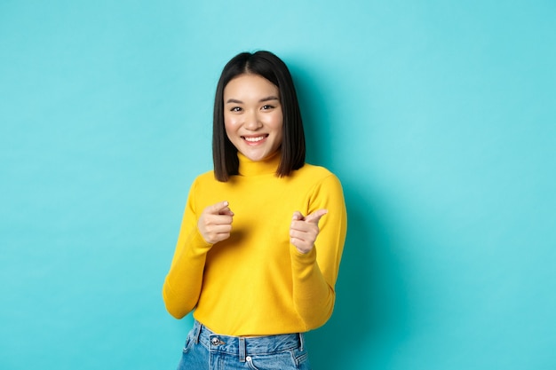 Sonriente joven mujer asiática señalando con el dedo a la cámara, eligiéndote, invitando al evento, feliz de pie contra el fondo azul.