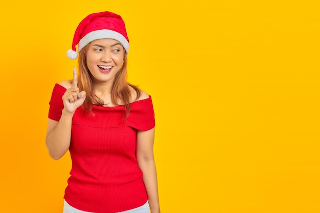 Sonriente joven mujer asiática con sombrero de Navidad apuntando con el dedo hacia arriba y tener una idea