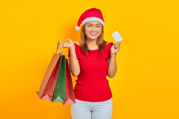 Foto sonriente joven mujer asiática con sombrero de santa claus sosteniendo bolsas de la compra y mostrando la tarjeta en blanco