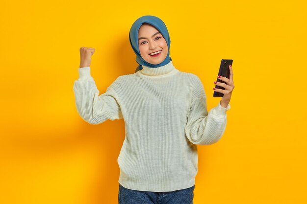 Sonriente joven mujer asiática en suéter blanco y hiyab usando teléfono móvil haciendo gesto de ganador aislado sobre fondo amarillo Concepto religioso del islam de la gente