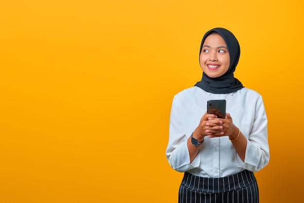 Sonriente joven mujer asiática con un teléfono móvil y mirando hacia los lados sobre fondo amarillo