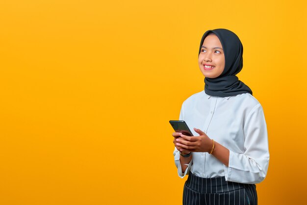 Sonriente joven mujer asiática con un teléfono móvil y mirando hacia los lados sobre fondo amarillo