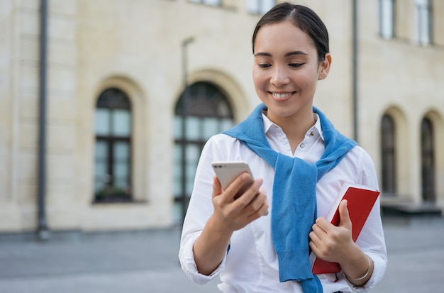 Sonriente joven mujer asiática mediante teléfono móvil, de pie en la calle