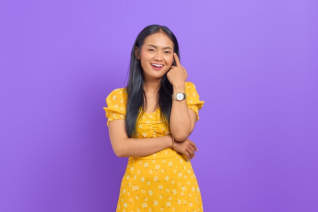 Sonriente joven mujer asiática con vestido amarillo y mirando confiado sobre fondo púrpura