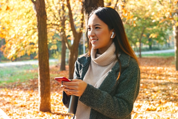 Sonriente joven mujer asiática vistiendo abrigo caminando al aire libre en el parque, escuchando música con auriculares, sosteniendo el teléfono móvil