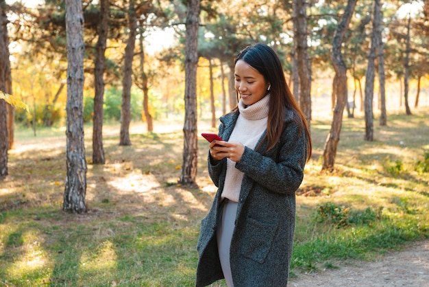 Sonriente joven mujer asiática vistiendo abrigo caminando al aire libre en el parque, escuchando música con auriculares, sosteniendo el teléfono móvil