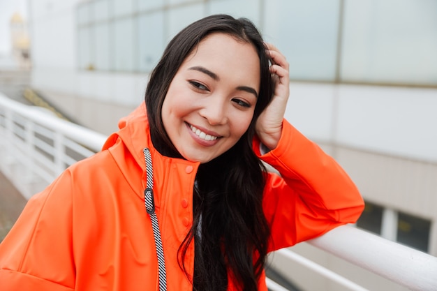 Sonriente joven mujer asiática vistiendo impermeable caminar al aire libre bajo la lluvia