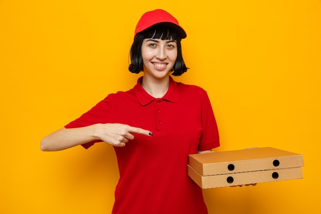 Sonriente joven mujer caucásica de entrega sosteniendo y apuntando a cajas de pizza