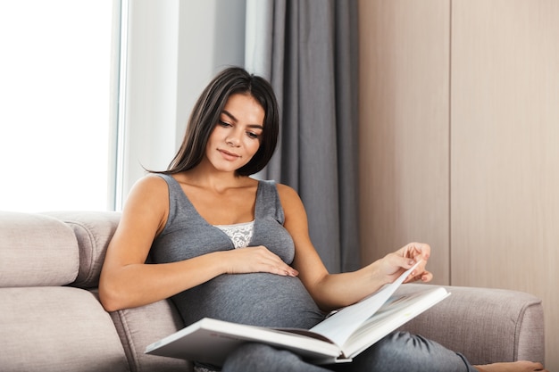 Sonriente joven mujer embarazada en casa, sentada en un sofá, leyendo libros