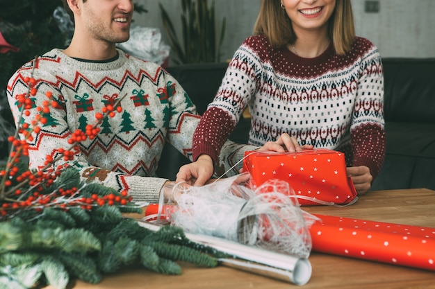 Sonriente joven y mujer envolver un regalo de Navidad vistiendo suéteres de Navidad en casa