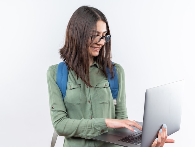 Sonriente joven mujer de la escuela con gafas con mochila sosteniendo y portátil usado