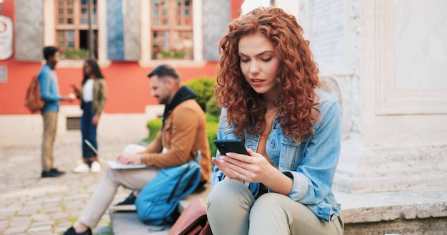 Foto sonriente joven mujer de jengibre con ropa casual con gadget de teléfono inteligente al aire libre en la calle whi ...