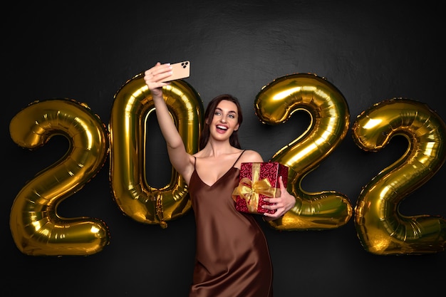 Sonriente joven mujer morena en vestido elegante haciendo selfie en teléfono inteligente sobre fondo negro dorado ...