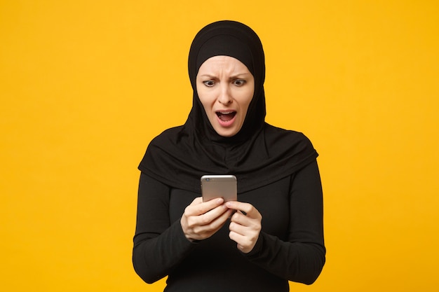 Sonriente joven mujer musulmana árabe en ropa negra hijab mantenga en manos teléfono móvil, charlando aislado en retrato de pared amarilla. Concepto de estilo de vida religioso de la gente. .