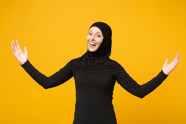 Sonriente joven mujer musulmana árabe en ropa negra hijab que muestra el espacio de la copia señalando con el dedo de las manos aislado en el retrato de la pared amarilla. Concepto de estilo de vida religioso de la gente.