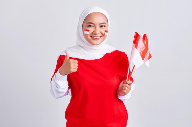 Sonriente joven mujer musulmana asiática en camiseta blanca roja sosteniendo la bandera indonesia para celebrar el día de la independencia de Indonesia el 17 de agosto y mostrando gesto de pulgar hacia arriba aislado en fondo blanco