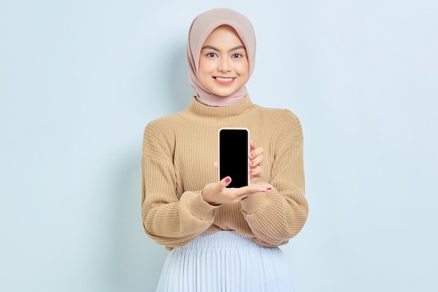 Sonriente joven mujer musulmana asiática en suéter marrón mostrando teléfono móvil de pantalla en blanco recomendando aplicación móvil aislada sobre fondo blanco Mockup teléfono celular de pantalla en blanco