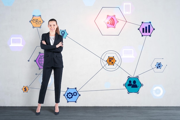 Sonriente joven mujer de negocios con traje negro de pie con los brazos cruzados cerca de una pared de hormigón con coloridos íconos de Internet dibujados en ella. Concepto de alta tecnología en los negocios.