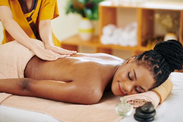 Sonriente joven mujer negra recibiendo relajante masaje de espalda en salón de belleza con aceites aromáticos