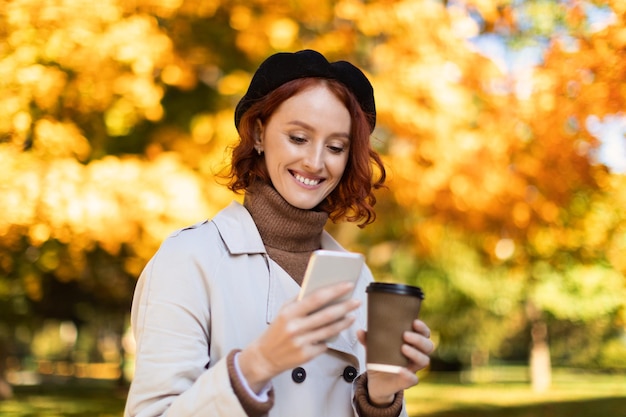 Sonriente joven mujer pelirroja europea con impermeable y había leído un mensaje en el teléfono inteligente disfruta de una bebida para llevar