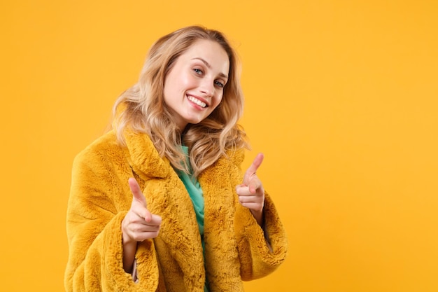 Sonriente joven mujer rubia chica en abrigo de piel amarillo posando aislado en retrato de estudio de fondo de pared naranja. Concepto de estilo de vida de las emociones de las personas. Simulacros de espacio de copia. Señalando con el dedo índice a la cámara.