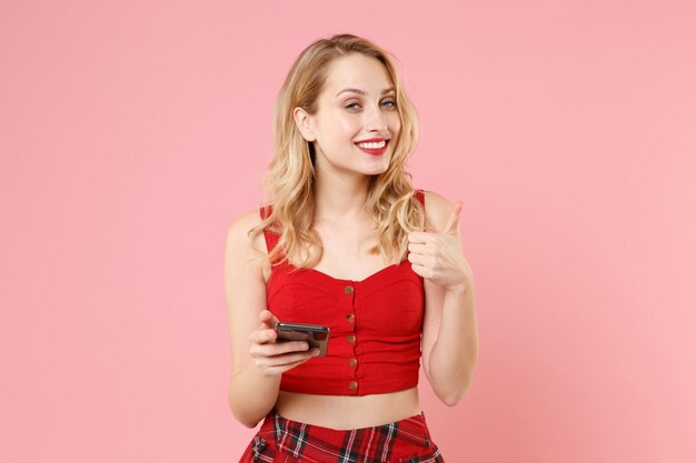 Sonriente joven mujer rubia chica en ropa sexy roja posando aislada sobre fondo rosa pastel. Concepto de estilo de vida de las personas. Simulacros de espacio de copia. Usando el teléfono móvil, escribiendo un mensaje sms, mostrando el pulgar hacia arriba.