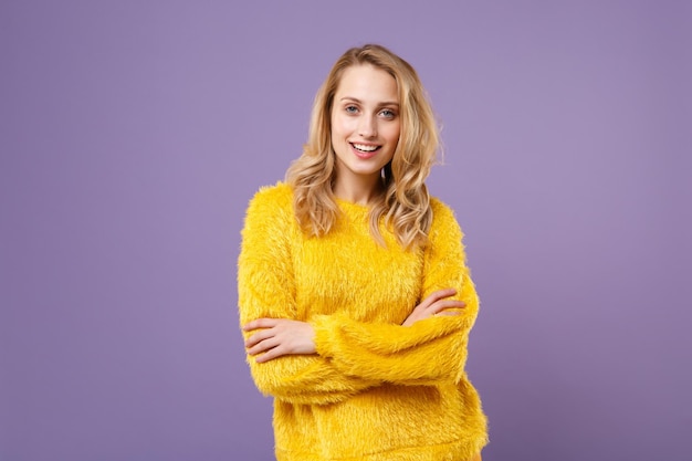 Sonriente a una joven mujer con suéter de piel amarilla posando aislada en un retrato de estudio de fondo violeta pastel. Gente emociones sinceras concepto de estilo de vida. Simulacros de espacio de copia. Tomados de la mano cruzados.