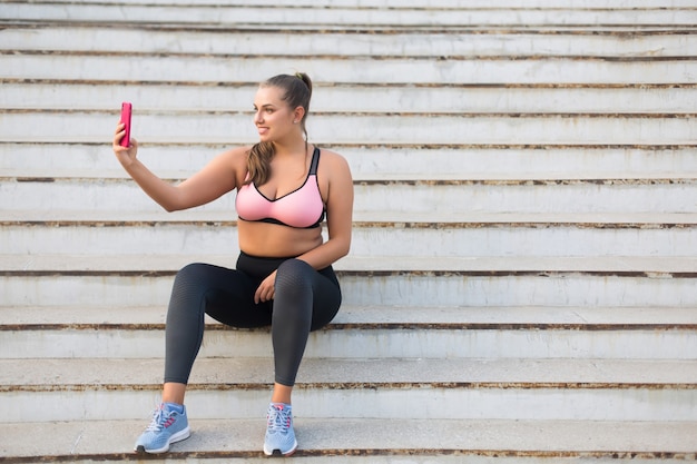 Sonriente joven mujer de talla grande en top deportivo y leggings sentado en las escaleras felizmente tomando fotos en el teléfono celular mientras pasa tiempo al aire libre