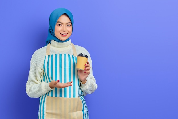 Sonriente joven musulmana asiática de unos 20 años con hiyab y delantal que muestra una taza de café de papel con las manos aisladas sobre un fondo morado Gente ama de casa concepto de estilo de vida musulmán