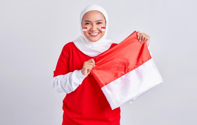 Sonriente joven musulmana asiática con camiseta blanca roja celebrando el día de la independencia de Indonesia el 17 de agosto con orgullo de mostrar la bandera indonesia aislada de fondo blanco