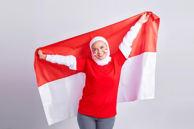Sonriente joven musulmana asiática con camiseta blanca roja celebrando el día de la independencia de Indonesia con orgullo mostrando la bandera de Indonesia aislada en el fondo blanco