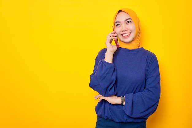 Sonriente joven musulmana asiática vestida con suéter casual hablando por teléfono móvil con espacio de copia aislado sobre fondo amarillo
