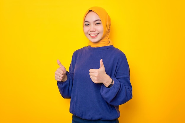 Sonriente joven musulmana asiática vestida con un suéter informal que muestra un gesto de aprobación con la mano recomendando algo bueno aislado sobre un fondo amarillo