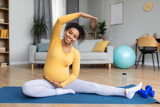 Sonriente joven negra embarazada con gran barriga haciendo ejercicios practicando yoga estirando la mano