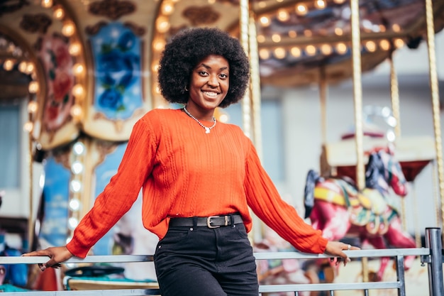 Sonriente joven negra con peinado afro y suéter rojo al lado de un carrusel en la calle