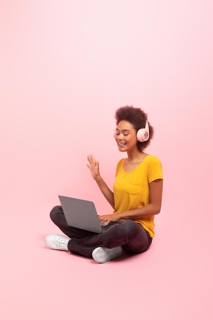 Sonriente joven negra rizada con auriculares inalámbricos sentada en el suelo con la computadora saludando con la mano