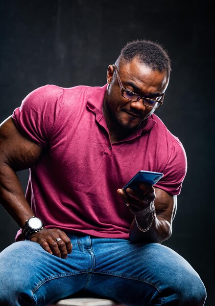 Sonriente joven negro alegre posa con el teléfono en las manos sobre fondo negro afroamericano en camisa rosa