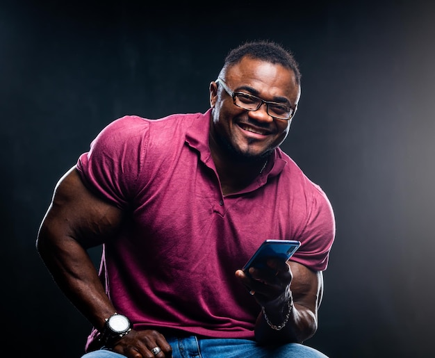 Sonriente joven negro alegre posa con el teléfono en las manos sobre fondo negro afroamericano en camisa rosa