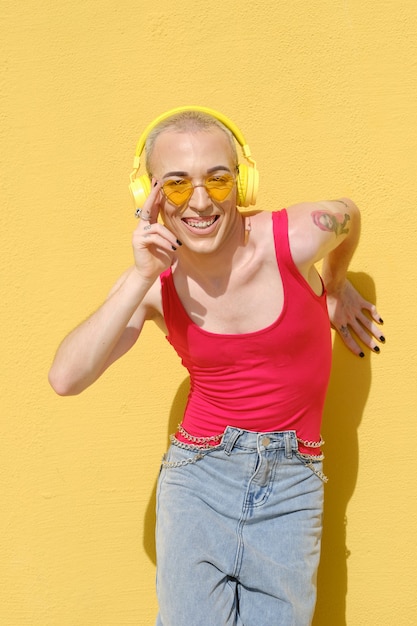 Foto sonriente joven no binario escuchando música con auriculares inalámbricos amarillos al aire libre contra una pared amarilla. concepto de tecnología y estilo de vida urbano.