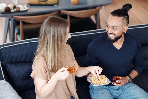 Sonriente joven pareja bebiendo té con galletas y discutiendo noticias y chismes en casa