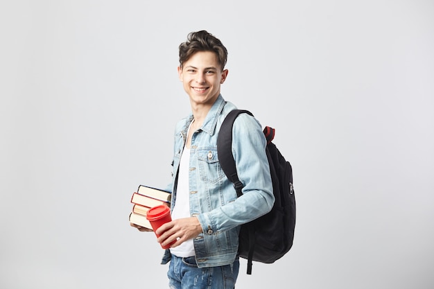 Sonriente joven de pelo oscuro sostiene libros y una taza de café en sus manos sobre el fondo blanco.