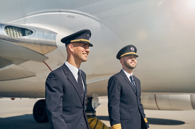 Sonriente joven piloto y su colega de pie junto a la aeronave aterrizaron en el aeródromo