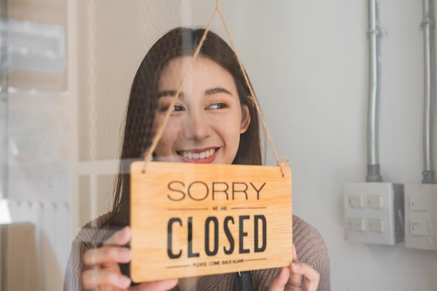 Foto sonriente joven propietaria asiática minorista, mujer de cafetería que cierra el letrero después de terminar el trabajo, el trabajo y la protección contra la pandemia de coronavirus. cerrar tienda debido a cierre, cuarentena de covid.