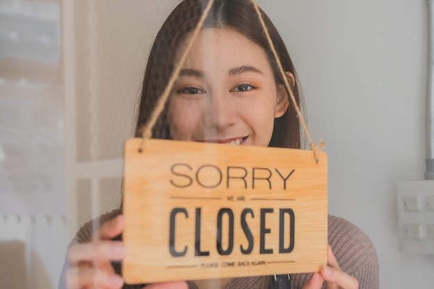 Foto sonriente joven propietaria de una cafetería minorista asiática que cambia el letrero a cerrado después de terminar el trabajo y la protección contra la pandemia de coronavirus cerrar la tienda debido a la cuarentena de bloqueo de covid