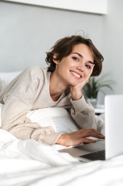 Sonriente joven relajante en la cama por la mañana, utilizando equipo portátil