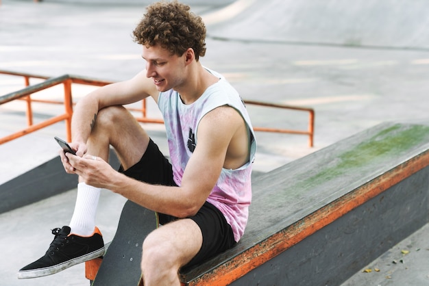 Sonriente joven en ropa de calle sentado con patineta y escribiendo en el teléfono celular en el parque de patinaje de verano