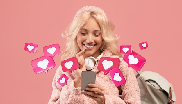 Sonriente joven rubia europea con mochila tiene chat romántico con corazones en el teléfono inteligente