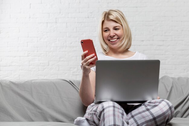 Sonriente joven rubia en ropa de casa está sentada en el sofá con una computadora y un teléfono. Trabajo a distancia y redes sociales. Espacio para texto.