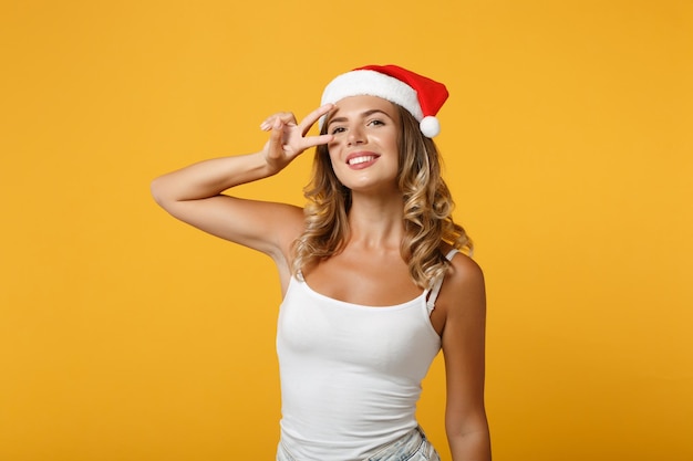 Sonriente joven Santa niña en camisa blanca, sombrero de Navidad posando aislado sobre fondo amarillo, retrato de estudio. Feliz Año Nuevo 2020 concepto de vacaciones de celebración. Simulacros de espacio de copia. Mostrando el signo de la victoria.